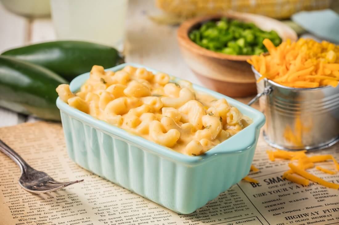 Image of a creamy Dave's Cheesy Mac & Cheese, served in a small, light turquoise casserole dish. Behind it sits two whole jalapenos, a ramekin of chopped green onions and shredded cheese in a small pail
