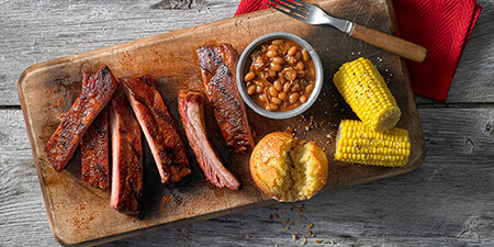 Image of five ribs cut up on a wooden cutting board, accompanied by a side of baked beans in a white bowl, corn bread, and two piece of corn on the cob