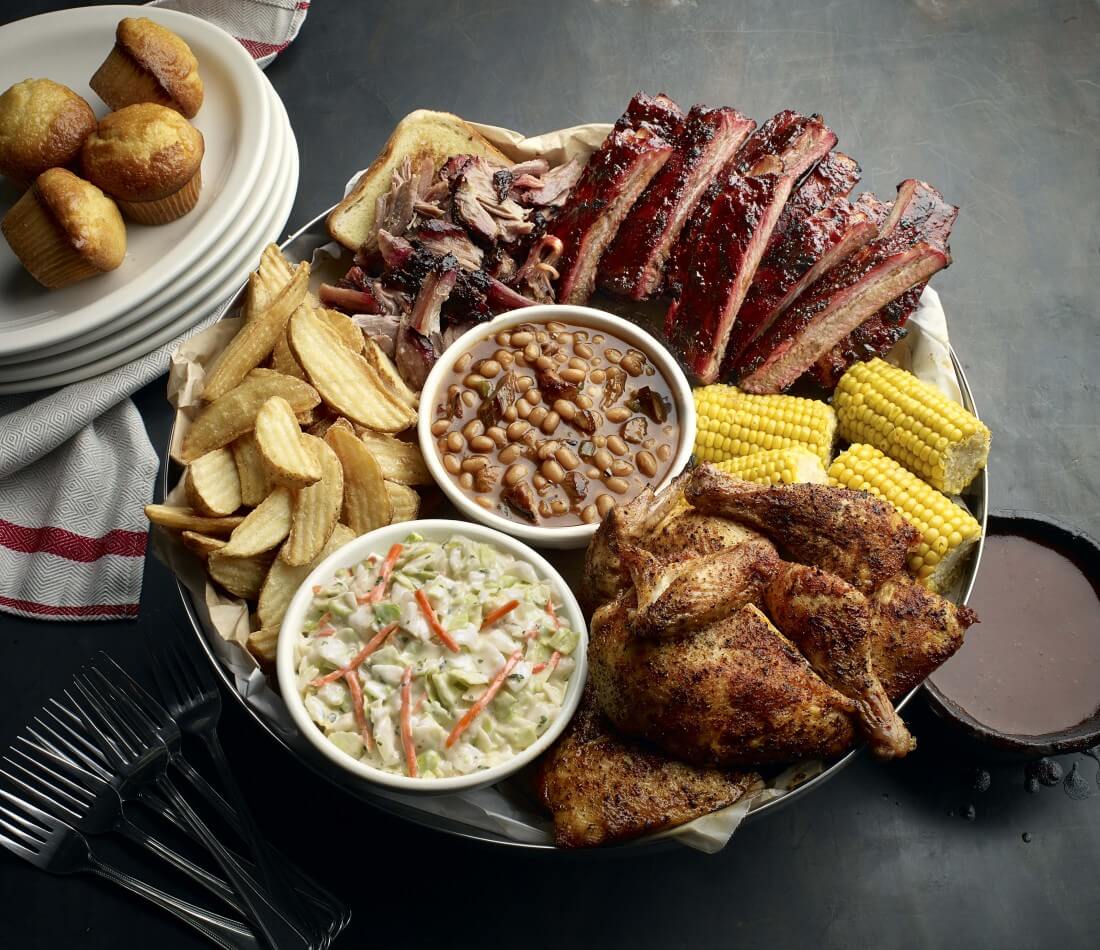 Famous Dave's BBQ Platter on a black tray with roasted chicken, chicken wings, breaded chicken, ribs, pickles, shoestring pickles, pickled onions, celery and carrot sticks, and various dipping sauces